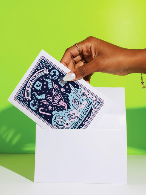 A vertical 5x7 holiday card featuring a retro tattoo style illustration of Krampus in pastel pink, teal, and black ink on shimmery silver pearlescent paper. The central horned demon has a long tongue and is surrounded by vintage rotary phones, daggers, candy canes, and skull gifts. Top text asks Have You Been Good while the bottom banner reads Krampus Is Calling. The interior text reads Let's be honest... Being good is overrated. Merry Christmas.