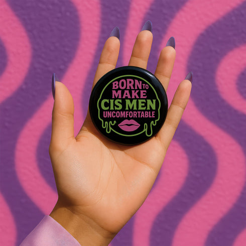 Glossy black pinback button featuring bold text 'Born to Make Cis Men Uncomfortable' in pink and green glitter fonts. Design includes melting skull outline and lips illustration. Button is round and 2.25 inches in diameter with a durable tinplate body and UV-resistant coating. Perfect for jackets, backpacks, or gifts for queer punks and feminists.