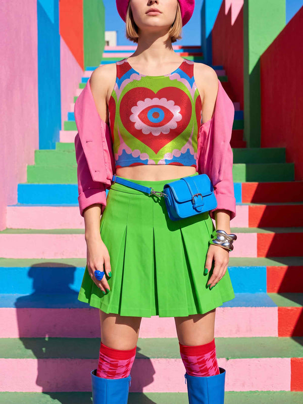 A sleeveless scoop neck crop top featuring a bold all-over printed design. The central graphic is a large bright red heart enclosing a scalloped pink flower shape and a blue evil eye motif in the middle. The background surrounds the heart with mirrored concentric wavy arches and scalloped borders in vibrant shades of lime green, pink, blue, and red. The fitted garment has a smooth athletic texture. Available in a standard matte performance knit or a sparkly silver glitter foil finish.