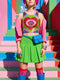 A sleeveless scoop neck crop top featuring a bold all-over printed design. The central graphic is a large bright red heart enclosing a scalloped pink flower shape and a blue evil eye motif in the middle. The background surrounds the heart with mirrored concentric wavy arches and scalloped borders in vibrant shades of lime green, pink, blue, and red. The fitted garment has a smooth athletic texture. Available in a standard matte performance knit or a sparkly silver glitter foil finish.
