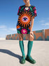 A short-sleeve, above-the-knee t-shirt dress with a straight, structured fit. The dress has a true black background featuring a bold, vibrant graphic pattern. The center displays a large blue and white evil eye surrounded by a bright orange wavy sunburst with a white jagged border. Above and below the eye are bright pink daisy flowers with orange centers, scattered white four-petal stars, and wavy blue and teal green leaf shapes. The fabric is thick, smooth, and holds its clean shape.
