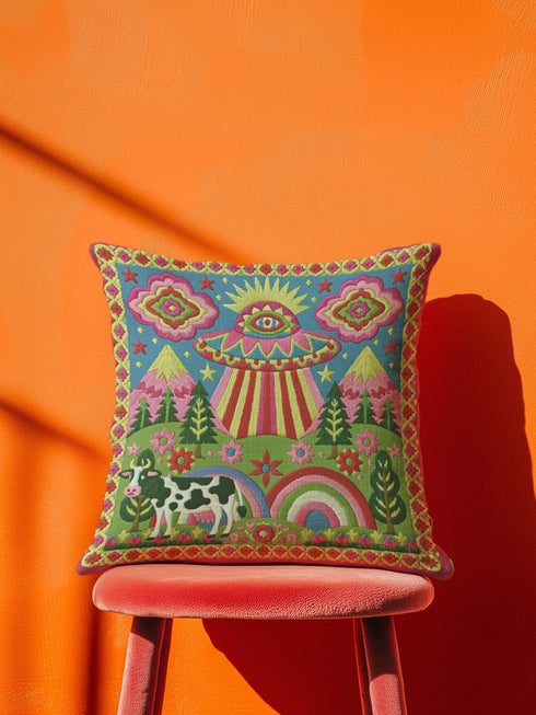 Description of a square throw pillow with a vibrant maximalist alien design. A central UFO with a large all-seeing eye hovers above a landscape with two rainbows, pink mountains, and green pine trees. A black and white spotted cow stands in the foreground. The design is a high-contrast palette of sky blue, magenta, hot pink, and meadow green. The pillow has a faux-embroidery 2D print finish on linen-feel fabric, framed by a yellow and pink scalloped floral border.