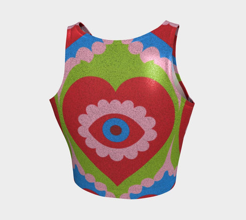 A sleeveless scoop neck crop top featuring a bold all-over printed design. The central graphic is a large bright red heart enclosing a scalloped pink flower shape and a blue evil eye motif in the middle. The background surrounds the heart with mirrored concentric wavy arches and scalloped borders in vibrant shades of lime green, pink, blue, and red. The fitted garment has a smooth athletic texture. Available in a standard matte performance knit or a sparkly silver glitter foil finish.