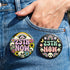 A round pinback button featuring a solid black background covered in a retro pastel colored design. The center reads 25TH NOW in a groovy bubbly font using pale pink and mint green referencing the 25th amendment. Surrounding the text is a maximalist cluster of cute but edgy motifs including smiling white skulls, pastel rainbows, pale yellow crescent moons, daisies, and a pink and white spotted mushroom. The button has a glossy laminate finish and a secure metal safety pin backing.