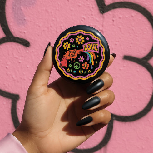 Round pinback button badge with a black background, featuring a retro illustration of a red revolver shooting out a rainbow flag that says “LOVE.” Surrounded by colorful 70s-style flowers and peace signs, outlined in wavy neon border. Glossy finish with vibrant pop colors and psychedelic design.