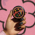 Round pinback button badge with a black background, featuring a retro illustration of a red revolver shooting out a rainbow flag that says “LOVE.” Surrounded by colorful 70s-style flowers and peace signs, outlined in wavy neon border. Glossy finish with vibrant pop colors and psychedelic design.