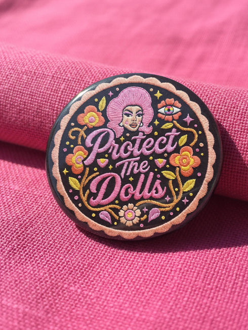 A circular black pinback button featuring the slogan Protect The Dolls in retro script. The design utilizes a high-fidelity trompe-l'œil print style that mimics the texture of raised thread embroidery with satin-stitch detailing. The artwork features vibrant hot pink, rust orange, and golden yellow motifs including drag queens with bouffant hair, 70s style daisies, lightning bolts, and a crescent moon with a face. The surface is smooth and glossy, creating a faux-textured illusion.