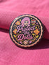 A circular black pinback button featuring the slogan Protect The Dolls in retro script. The design utilizes a high-fidelity trompe-l'œil print style that mimics the texture of raised thread embroidery with satin-stitch detailing. The artwork features vibrant hot pink, rust orange, and golden yellow motifs including drag queens with bouffant hair, 70s style daisies, lightning bolts, and a crescent moon with a face. The surface is smooth and glossy, creating a faux-textured illusion.
