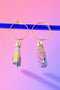 A pair of gold hoop earrings with colorful gemstones hanging from them against a gradient pink and purple background.