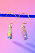 A pair of gold hoop earrings with colorful gemstones hanging from them against a gradient pink and purple background.