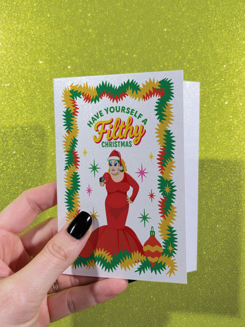 A 5x7 pearlescent holiday card by HoneyDazed. The card illustration, inspired by John Waters, features the drag queen Divine in a red dress with a Santa hat. She points a gun, withtext reads "Have Yourself a Filthy Christmas" in campy font. The card is bordered by garland. Iinside of the card reads: "Season's Sleazings" in red and green text. This kitschy greeting card is a perfect piece of cult film memorabilia and a funny adult humor gift for fans of queer art.