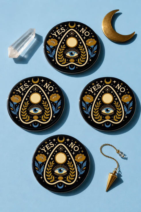 A close-up photograph of a glossy ceramic coaster featuring a Planchette occult design on a deep black background. The symmetrical graphic displays a central blue All-Seeing Eye with gold sunburst lines, topped by a white circle, and flanked by gold flowers with blue stems. At the top, the text "YES" is printed in gold on the left and "NO" on the right. Gold stars and crescent moons are scattered around the perimeter. The flat, round coaster has a highly visible high-gloss glaze.