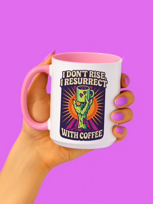 White ceramic coffee mug with red handle and interior, featuring a bold design of a zombie hand holding a coffee cup with the text “I Don’t Rise, I Resurrect With Coffee” in white and purple, accented with a neon green hand and orange sunburst background.