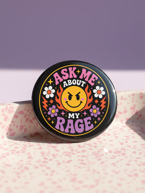 A round glossy metal pinback button featuring a flat 2D print design on a solid black background. In the center is an angry yellow smiley face surrounded by orange and red flames. Above the smiley face, pink text reads ASK ME and white text reads ABOUT. Below the face, white text reads MY and large purple text reads RAGE. The design is accented with two white daisies, two purple flowers, pink and orange four-point stars, and orange lightning bolts framing the outer edges.