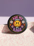 A round glossy metal pinback button featuring a flat 2D print design on a solid black background. In the center is an angry yellow smiley face surrounded by orange and red flames. Above the smiley face, pink text reads ASK ME and white text reads ABOUT. Below the face, white text reads MY and large purple text reads RAGE. The design is accented with two white daisies, two purple flowers, pink and orange four-point stars, and orange lightning bolts framing the outer edges.