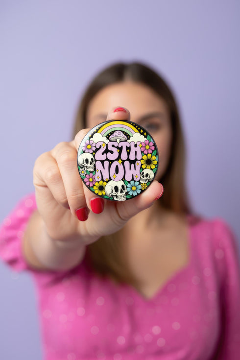 A round pinback button featuring a solid black background covered in a retro pastel colored design. The center reads 25TH NOW in a groovy bubbly font using pale pink and mint green referencing the 25th amendment. Surrounding the text is a maximalist cluster of cute but edgy motifs including smiling white skulls, pastel rainbows, pale yellow crescent moons, daisies, and a pink and white spotted mushroom. The button has a glossy laminate finish and a secure metal safety pin backing.