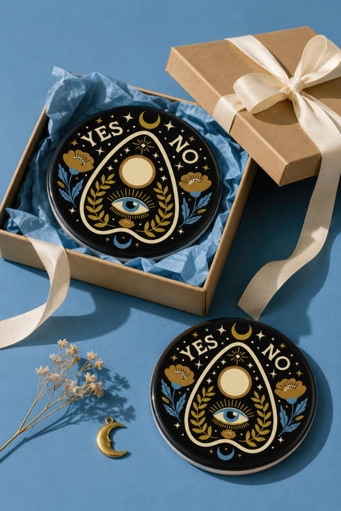 A close-up photograph of a glossy ceramic coaster featuring a Planchette occult design on a deep black background. The symmetrical graphic displays a central blue All-Seeing Eye with gold sunburst lines, topped by a white circle, and flanked by gold flowers with blue stems. At the top, the text "YES" is printed in gold on the left and "NO" on the right. Gold stars and crescent moons are scattered around the perimeter. The flat, round coaster has a highly visible high-gloss glaze.