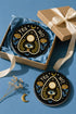 A close-up photograph of a glossy ceramic coaster featuring a Planchette occult design on a deep black background. The symmetrical graphic displays a central blue All-Seeing Eye with gold sunburst lines, topped by a white circle, and flanked by gold flowers with blue stems. At the top, the text "YES" is printed in gold on the left and "NO" on the right. Gold stars and crescent moons are scattered around the perimeter. The flat, round coaster has a highly visible high-gloss glaze.