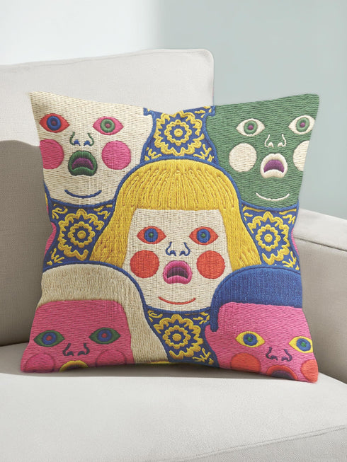 A square decorative pillow cover featuring a faux embroidery pattern of five stylized, expressive doll faces. The central face has yellow hair and pink cheeks. It is surrounded by four other faces in shades of pale beige, green, and bright pink, all with wide staring eyes and open mouths. The design uses intricate, colorful line work to create the illusion of thick woven stitching, but the fabric is a smooth print. Medallions fill the gaps.