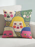 A square decorative pillow cover featuring a faux embroidery pattern of five stylized, expressive doll faces. The central face has yellow hair and pink cheeks. It is surrounded by four other faces in shades of pale beige, green, and bright pink, all with wide staring eyes and open mouths. The design uses intricate, colorful line work to create the illusion of thick woven stitching, but the fabric is a smooth print. Medallions fill the gaps.
