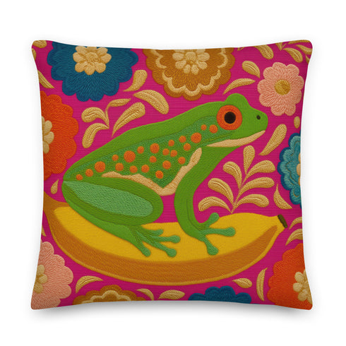 A square decorative throw pillow made from premium polyester with a visible linen-like woven texture. The background is a vibrant hot pink covered in a dense, retro-style floral pattern in orange, teal, and gold. The main design is a brightly colored, stylized illustration of a green red-eyed tree frog with orange feet and prominent red eyes. The whimsical frog is riding a vibrant yellow banana. The overall aesthetic is colorful, maximalist, and eclectic, with a faux-embroidered texture effect.