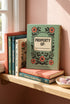A sage green hardcover spiral planner for 2027-2028 with the title "The 2027 Planner of Controlled Chaos" in a funky, retro 70s-style font. The cover features a groovy illustration of orange and pink flowers in a vase, with one flower depicted as an eyeball. The text reads "Bloom where you're planted. Or just wilt artfully." and "Notes from the Void." The planner has a gold metal wire-o binding. The back cover shows a decorative floral frame with a "Property Of" section