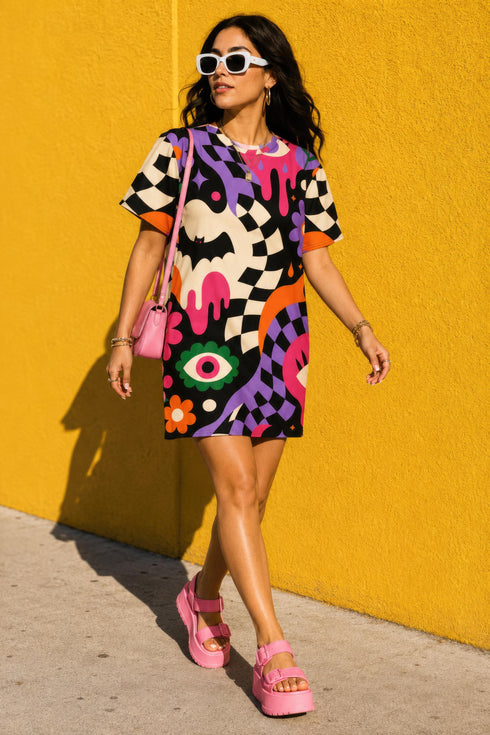 A short-sleeve, above-the-knee t-shirt dress with a relaxed, straight fit and a crew neckline. The dress features a vibrant, chaotic, all-over graphic print. The design includes sections of distorted, wavy black and cream checkerboard, mixed with bold, abstract shapes in bright purple, hot pink, and vivid orange. Scattered throughout the pattern are motifs of hot pink dripping slime, a black bat silhouette, a stylized green eye with a pink iris, and retro pink and purple daisy flowers.