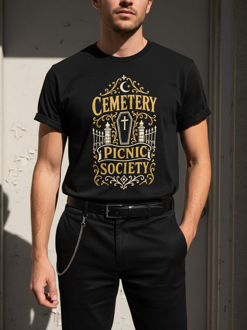 A black unisex crewneck graphic t-shirt featuring an ornate Victorian-inspired emblem printed on the chest in vintage gold and off-white. The centralized, symmetrical design showcases a classic coffin with a cross on the lid, flanked by wrought-iron cemetery gates. Beautiful scrollwork and decorative filigree flourishes surround the bold, retro serif text reading CEMETERY PICNIC SOCIETY. A crescent moon and stars sit at the top, completing this gothic memento mori graphic shirt.
