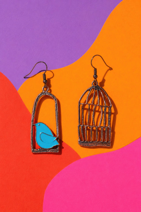 two earrings, one with a bird inside a cage and the other with a bird outside the cage, set against a vibrant background of purple, orange, and pink.