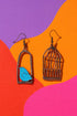 two earrings, one with a bird inside a cage and the other with a bird outside the cage, set against a vibrant background of purple, orange, and pink.