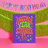 A birthday card with the text "Another Year Stranger" is displayed in front of a pink background with a banner that says "Happy Birthday".