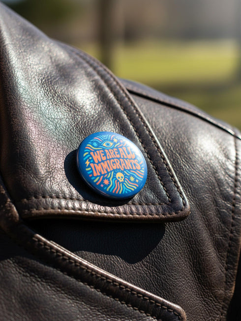 A round 1.75 inch blue pinback button featuring retro psychedelic artwork. The text reads We Are All Immigrants in a groovy orange and red 1970s font style centered on the badge. A mystic third eye sits above the text while a small skull sits below it. Wavy rainbow lines in cyan, green, and orange radiate outward from the center along with small starbursts and lightning bolts. The button has a glossy finish reflecting light.