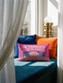 A bright magenta pink rectangular lumbar pillow cover featuring a flat 2D printed faux embroidery design. The printed illusion shows light pink bubble text reading NO THOUGHTS JUST BLANKETS. Below the text is a cartoon pink brain with a happy face, tucked into a blue bed with blue pillows. The brain is covered by a textured-looking orange blanket decorated with light purple daisies. The entire design is a smooth printed trompe l'oeil stitch effect, not real woven or stitched texture.