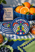 A close-up photograph of a round, glossy ceramic coaster featuring a bold retro pop art design. The background is a deep royal blue. In the center are two symmetrical profile faces of twins looking at each other, featuring bright green skin, closed eyes, and red cheeks. They have wavy white and pale pink hair with red accents. The word Gemini is arched at the top in a retro white font. The design is decorated with a green Gemini symbol, pink and red hearts, and white starbursts.