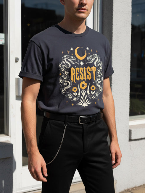 A unisex short sleeve graphic t shirt in dark charcoal black featuring a highly detailed symmetrical medallion print on the chest. In the center, the word RESIST is printed in a bold yellow gothic blackletter font. Two coiled white snakes with fine black scale details frame the text on the left and right. Above the text, a yellow crescent moon is flanked by white and yellow starbursts. Beneath, three yellow marigold flowers sit atop white stems and leafy foliage.