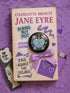 Glossy black pinback button featuring embroidered-style artwork of a pastel purple heart that reads “Read or Die” pierced by a sword and surrounded by roses; shown on books, graffiti, and lace backgrounds.