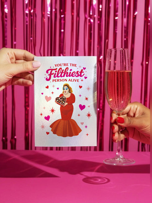 A 5x7 inch pearlescent silver greeting card featuring a flat vector illustration of the drag queen Divine in a red mermaid gown holding a heart-shaped box of chocolates. The text above reads "You're The Filthiest Person Alive" in bold red script with pink sparkles. The card has a shimmering finish that reflects light. Inside text reads "...and I love it." The back features the Honey Dazed logo. Includes a matching envelope.