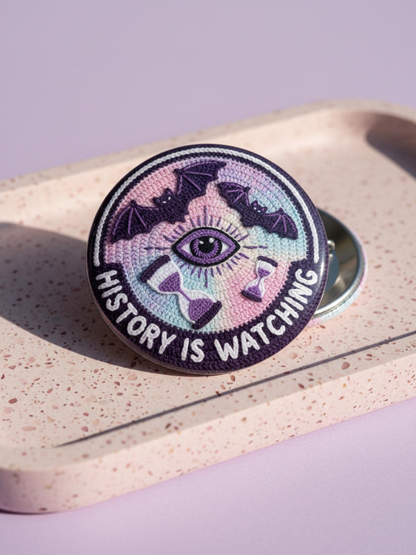 Round pinback button featuring a trompe-l'œil print mimicking 3D embroidery texture. Background is a pastel tie-dye gradient of pink, teal, and lavender. A central open eye with a purple iris sits below three flying plum bats and above two hourglasses with purple sand. A dark purple bottom rocker displays white text "HISTORY IS WATCHING." The flat glossy metal surface creates a realistic optical illusion of stitched fabric and raised threads.
