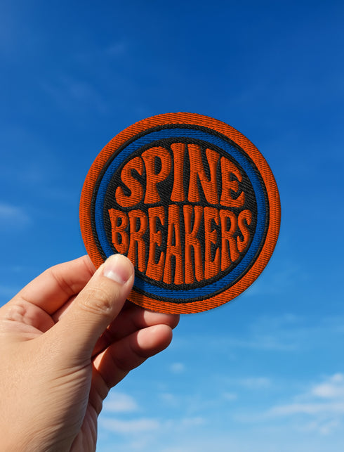 3-inch round embroidered iron-on patch made from a cotton–polyester blend; navy center with blue ring and orange outer ring; stacked orange text reads “SPINE BREAKERS”; merrowed border. Book club patch for paperback addicts, library tote flair, and literary merch collectors; makes a great gift for readers who annotate, dog-ear, and devour novels.