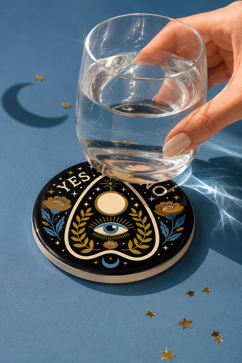 A close-up photograph of a glossy ceramic coaster featuring a Planchette occult design on a deep black background. The symmetrical graphic displays a central blue All-Seeing Eye with gold sunburst lines, topped by a white circle, and flanked by gold flowers with blue stems. At the top, the text "YES" is printed in gold on the left and "NO" on the right. Gold stars and crescent moons are scattered around the perimeter. The flat, round coaster has a highly visible high-gloss glaze.