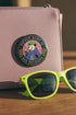 A round glossy metal pinback button featuring a flat 2D print illustration of an opossum wearing a blue cloak and a green crown. The opossum holds a neon green goblet in one paw and two purple dice in the other. A bright pink circle sits behind the opossum, surrounded by a black outer ring. Purple text reads PATRON SAINT OF BAD DECISIONS curved around the edge. Two pink roses with green leaves are on the sides. The design uses bold black outlines and bright neon colors.