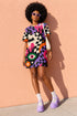 A short-sleeve, above-the-knee t-shirt dress with a relaxed, straight fit and a crew neckline. The dress features a vibrant, chaotic, all-over graphic print. The design includes sections of distorted, wavy black and cream checkerboard, mixed with bold, abstract shapes in bright purple, hot pink, and vivid orange. Scattered throughout the pattern are motifs of hot pink dripping slime, a black bat silhouette, a stylized green eye with a pink iris, and retro pink and purple daisy flowers.