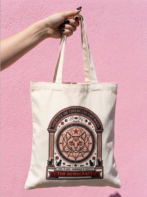 A beige canvas tote bag featuring a vintage-style circular crest design in rust orange and black. The text reads "Society of Childless Cat Dads" in an arch over a central illustration of a tabby cat face inside a geometric star and hexagon symbol. Below the cat, the text reads "Loyal to Cats, Suspicious of Fascists." A banner at the bottom reads "For Democracy." The design features occult and whimsigoth elements like crescent moons, columns, and fish bones.