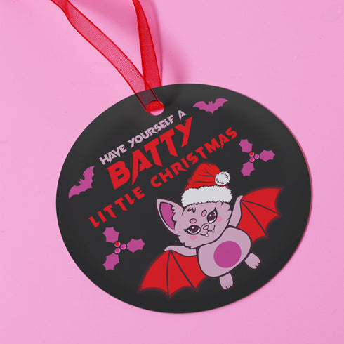 Round black metal Christmas ornament featuring cute pink bat with red wings wearing Santa hat, surrounded by pink bats and holly. Text reads "Have Yourself A Batty Little Christmas"