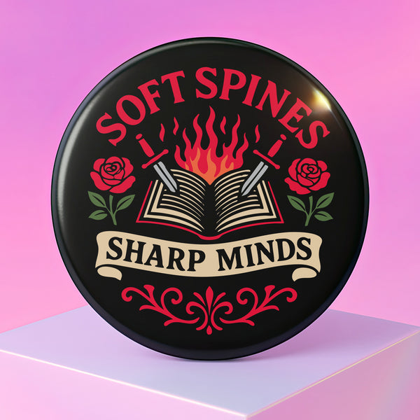 A round pinback button in black with a glossy mylar face and metal construction. The design, in a traditional tattoo style, features a central open book with flames rising from its pages. Two silver swords with black hilts are crossed over the book. On either side of the book is a red rose with a green stem and leaves. Below the book is a cream-colored banner with the words "SHARP MINDS" in a black serif font. Above the book, the words "SOFT SPINES" are written in a curved, red font.