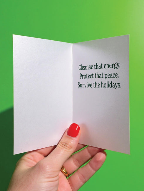 A detailed folk-art style illustration of a "Sage the Halls" holiday greeting card. The design features a bundle of sage smudge sticks tied with a red ribbon at the center of a dark green wreath. The wreath is accented with small crescent moons, stars, and smoke swirls. Below the wreath are two crossed matches with red tips. The card is printed on creamy beige paper with bold, dark green serif text. This funny spiritual and witchy Yule card is a unique pagan greeting for energy cleansing.