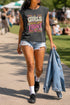 A women's garment-dyed muscle tee in faded charcoal grey, featuring a boxy, slightly cropped fit with structured shoulders and a wide ribbed collar. The center chest displays a retro graphic reading GIRLS TO THE FRONT in cream and magenta bubbly typography. The text is surrounded by vintage motifs including white daisies, yellow and purple stars, wavy ribbon banners, pink and yellow checkerboard borders, and a bottom radial sunburst in orange and magenta. Made of one hundred percent cotton.