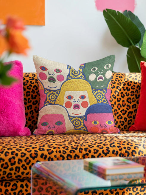A square decorative pillow cover featuring a faux embroidery pattern of five stylized, expressive doll faces. The central face has yellow hair and pink cheeks. It is surrounded by four other faces in shades of pale beige, green, and bright pink, all with wide staring eyes and open mouths. The design uses intricate, colorful line work to create the illusion of thick woven stitching, but the fabric is a smooth print. Medallions fill the gaps.