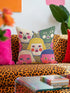 A square decorative pillow cover featuring a faux embroidery pattern of five stylized, expressive doll faces. The central face has yellow hair and pink cheeks. It is surrounded by four other faces in shades of pale beige, green, and bright pink, all with wide staring eyes and open mouths. The design uses intricate, colorful line work to create the illusion of thick woven stitching, but the fabric is a smooth print. Medallions fill the gaps.
