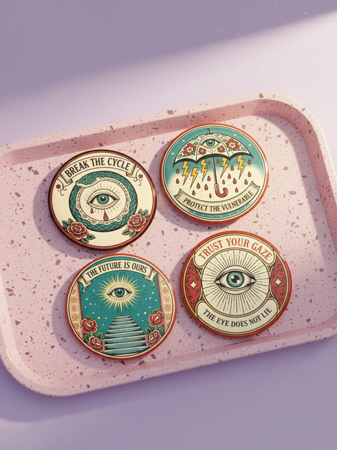 Four 2.25-inch circular metal pinback buttons on a pink textured background. The designs feature vintage tattoo-style art in rust, ochre, gold, and teal with heavy black linework. Designs include a snake Ouroboros "BREAK THE CYCLE," an umbrella eye "PROTECT THE VULNERABLE," a staircase "THE FUTURE IS OURS," and a radial eye "TRUST YOUR GAZE." The pins have a high-gloss finish. Perfect accessory for backpacks, jackets, and lanyards.